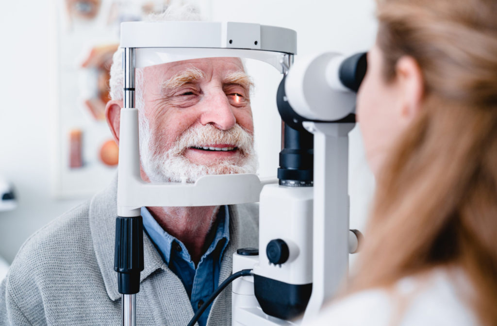 A male senior is sitting in front of a refracting machine and his chin is resting on chin rest while the optometrist is checking for refractive errors like myopia (nearsightedness), hyperopia (farsightedness), and astigmatism.