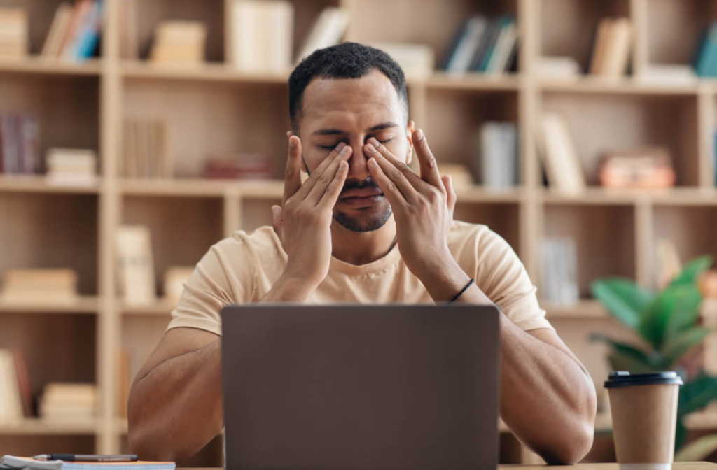 A fit young man rubbing his eyes with both hands while sitting in front of a laptop with a cup of coffee.