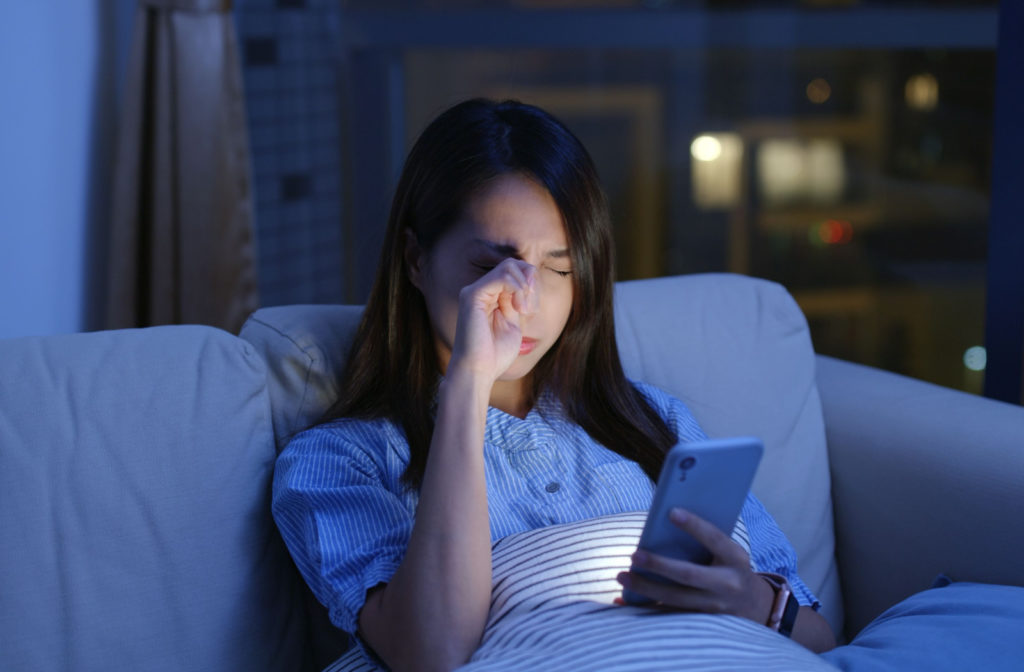 A woman rubbing her right eye with her right hand while using her phone in a dark room.