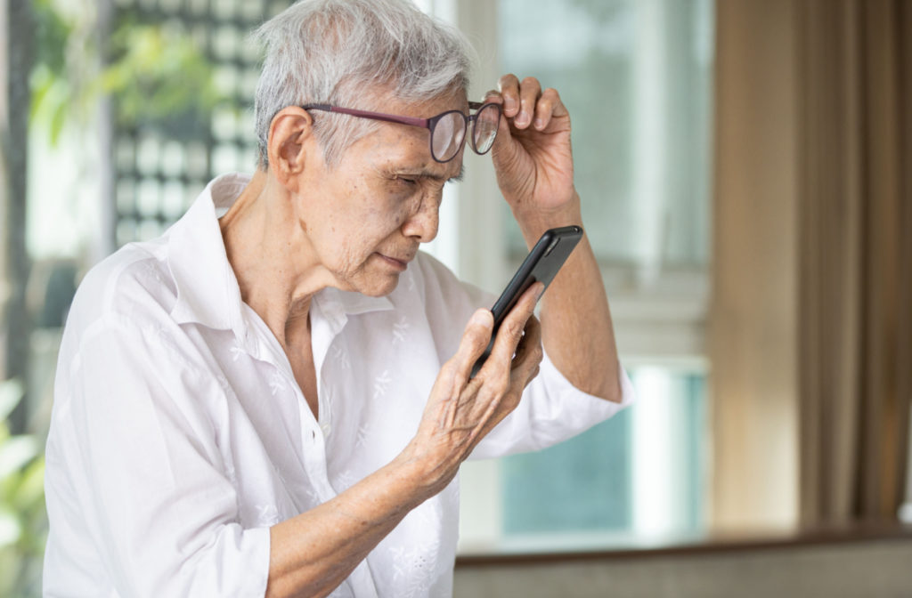 A senior woman holding her smartphone closer to her eyes while holding her eye glasses up to her forehead.