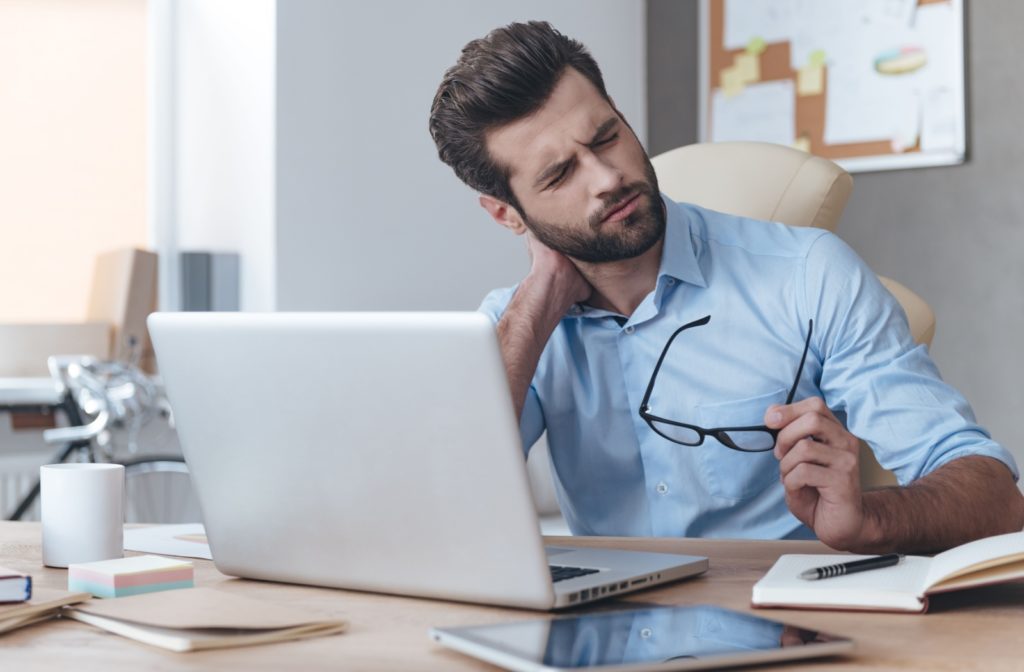 An exhausted office worker at a laptop attempts to rubs and stretch the tension from his neck.