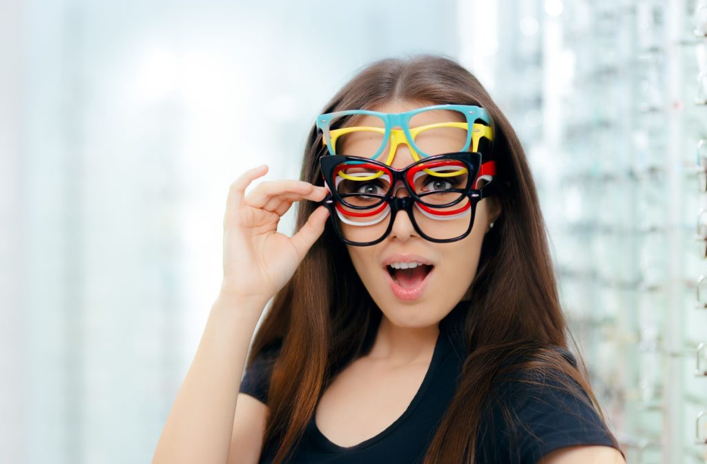 A woman with a playful expression and many pairs of eyeglasses frames in different colors on her face holding one pair with her right hand.
